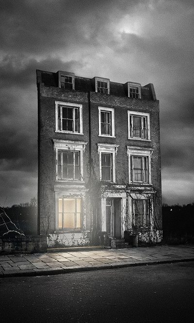 A tall, narrow, abandoned brick building stands alone at night under a cloudy sky. One lower window glows with light, whilst the rest appear dark and empty.