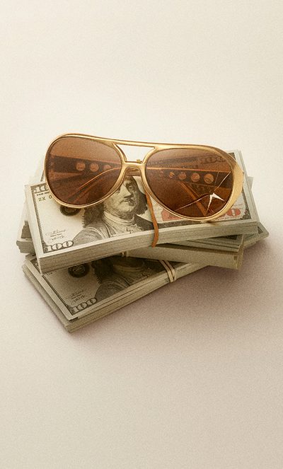 A pair of gold-framed sunglasses rests on top of several stacks of $100 U.S. bills, secured with rubber bands, on a plain, light surface.