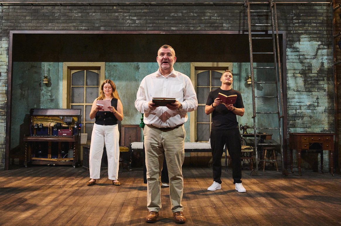 Three people stand on a theatre stage facing out holding books