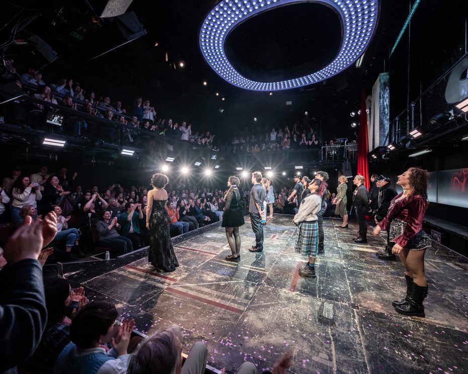 Actors stand on stage taking a bow as the audience are on their feet clapping enthusiastically