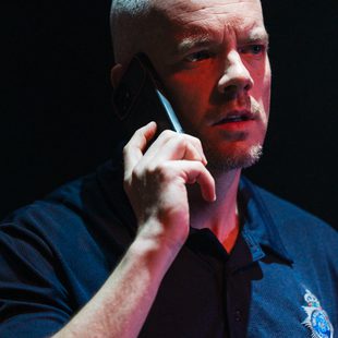 A man with a shaved head and short beard, wearing a dark polo shirt, holds a smartphone to his ear and looks concerned.