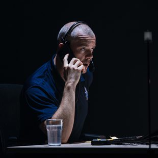 A man wearing a headset sits at a desk in a dimly lit room, talking on the phone and looking at a computer monitor. There is a glass of water and a keyboard on the desk.