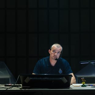 A man wearing a headset sits at a desk with a computer monitor, lamp, and small figurine, in a dimly lit room with covered furniture in the background. He looks directly ahead with a serious expression.
