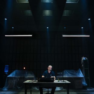 A person sits alone at a desk in a dimly lit, high-tech room with dramatic spotlights overhead. Covered equipment and cables are visible in the background, creating a mysterious, focussed atmosphere.