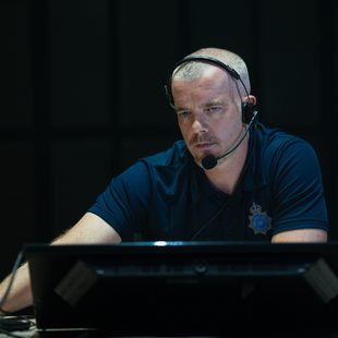 A man wearing a headset and a dark polo shirt with an official badge sits at a desk, looking intently at a computer monitor in a dimly lit room.