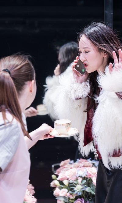 A woman is talking on a mobile phone. Another woman offers her a cup of tea but is ignored.