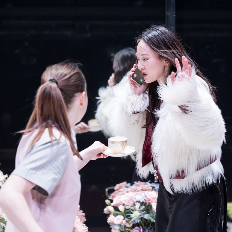 A woman is talking on a mobile phone. Another woman offers her a cup of tea but is ignored.