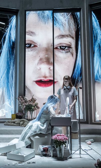 A woman in a blue wig looks into a mirror and her face is projected large on a screen behind her while another woman looks at her.
